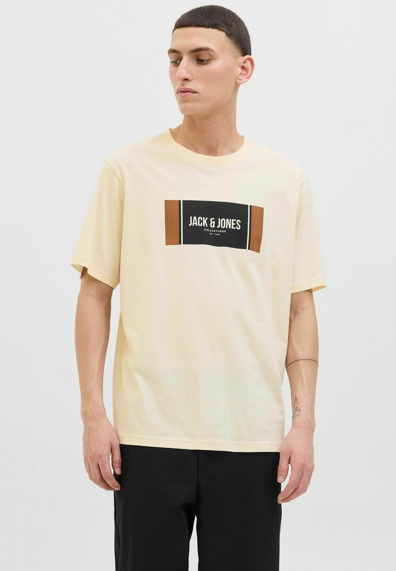 Noor mees, kes kannab beeži Jack & Jones T-särki musta ja pruuni logo plokiga, vaatab kõrvale, ning on kombo mustade pükstega.