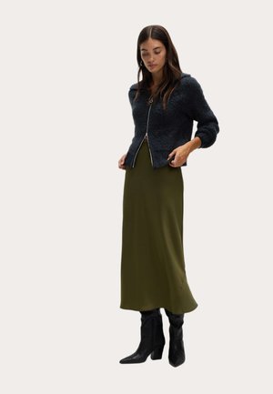 Cardigan de textura oscura con cremallera al frente, combinado con una falda larga verde oliva y botas de tacón alto negro con punta. 