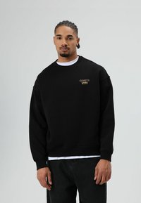 Zwarte trui met ronde hals, versierd met gouden borduursel waarop aan de linkerkant 'dreams Chasing' staat. Gladde stof, losse pasvorm.