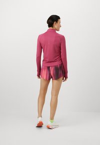 Fitted langärmliges pinkes Sporttop mit Daumenlöchern, kombiniert mit plissierten rosa und lila Shorts. Trägt weiße Schuhe mit neonfarbigen Akzenten.