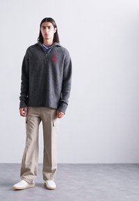 Grauer Reißverschluss-Pullover aus strukturiertem Stoff mit rotem Logo, kombiniert mit beigen Cargo-Hosen und weißen Sneakers. Schlichtes Design, lockere Passform.