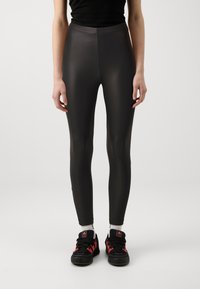 Leggings noirs, moulants, en matériau lisse et brillant, dotés d'une taille haute. Portés avec des chaussures de sport noires et rouges.