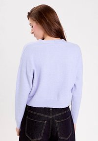 Kvinde med langt brunt hår iført en lys lilla cropped sweater og mørke højtaljede jeans, vendende væk mod en ensfarvet baggrund.