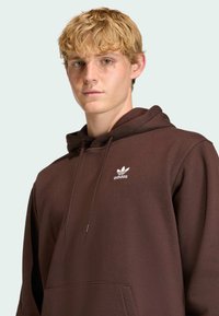 Hnedá mikina so soft touch materiálom, má klokanie vrecko a biele logo Adidas na ľavej hrudi. Obsahuje šnúrky na nastaviteľnú kapucňu.