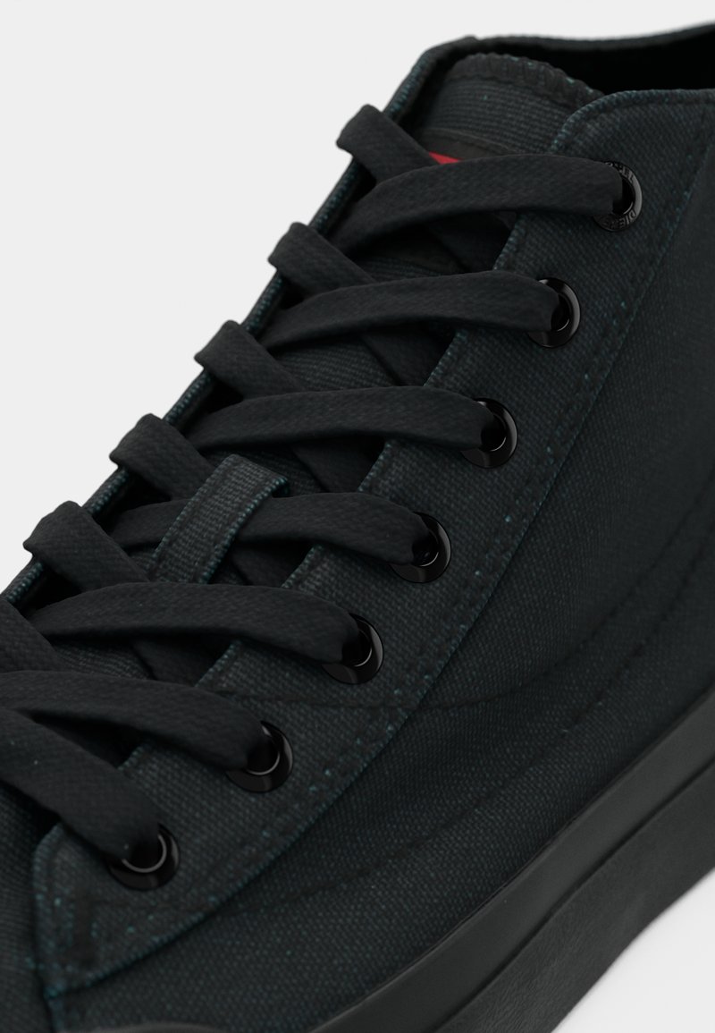 Schwarzer Canvas-Sneaker mit flachen schwarzen Schnürsenkeln, runden Ösen und kontrastierenden Nähten. Verfügt über eine strukturierte Oberfläche und eine Gummisohle.