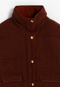 Veste matelassée de couleur rouille avec un col haut, ornée de boutons dorés et de deux poches avant avec des détails en rabat. Motif texturé sur l'ensemble.