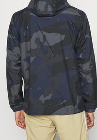 Vattentät marinblå kamouflagejacka med huva, elastiska muddar och en slät, texturerad yta med små vattendroppar.