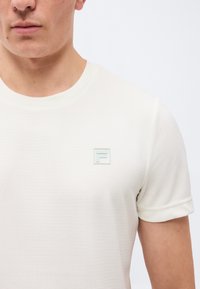 Άνδρας που φοράει απλό λευκό κοντομάνικο πουκάμισο με μικρό ανεβασμένο λογότυπο "F" στο αριστερό στήθος πάνω σε απλό φωτεινό φόντο.