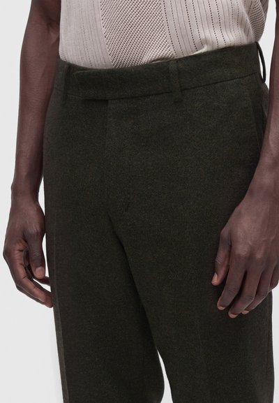 Close-up of a person wearing dark green wool trousers and a beige knit top, showing hands resting by their sides.