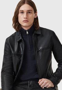 Schwarze Lederjacke mit einem Kragen-Design, vorderen Reißverschlusstaschen und gerippten Bündchen. Getragen über einem dunklen Sweater mit Reißverschluss am Hals. Glatte Textur.