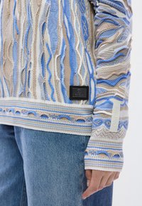 Πλεκτό πουλόβερ σε μπεζ και μπλε, με μοτίβο κύματος, ριμπ μανσέτες και λεπτομέρεια με μαύρη ετικέτα. Συνδυάζεται με ανοιχτό μπλε τζιν.
