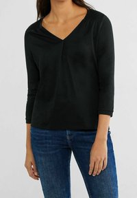 Svart topp med trekvartsärmar och V-ringning. Tillverkad av mjukt material, med diskret rynkning vid axlarna och en avslappnad passform.