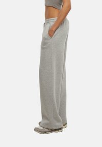 Pantaloni grigi in felpa realizzati in un tessuto morbido, con vestibilità ampia, tasche laterali e un logo ricamato sulla coscia. Abbinati a scarpe sportive.