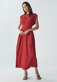 Červené šaty po kolena s páskem v pase, krátkými rukávy a strukturovaným materiálem. Oblečeno s černými sandály na pásky. Hladké pozadí.