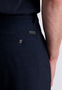 Nahaufnahme einer Person an der Taille, die dunkle, strukturierte Hose mit einer Gesäßtasche und einem kleinen Markenlabel trägt.