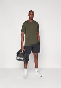 T-skjorte med korte ermer i olivengrønt, svarte shorts og hvite joggesko. Holder en svart duffelbag med hvit "adidas"-logo. Enkel, atletisk stil.