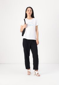 White T-shirt with blue text, black sweatpants, and open-toed white heels. Model holds a black shoulder bag. Simple design, casual style.