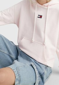 Světle růžová mikina s kapucí, přední kapsou, šňůrkami a malým černým a červeným logem. Spárována se světle modrými denimovými šortkami.