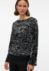 Črn zgornji del z bleščicami, ki ima okroglo izrezovanje, dolge rokave in teksturirano površino z odsevnimi bleščicami, v kombinaciji s črnimi hlačami.