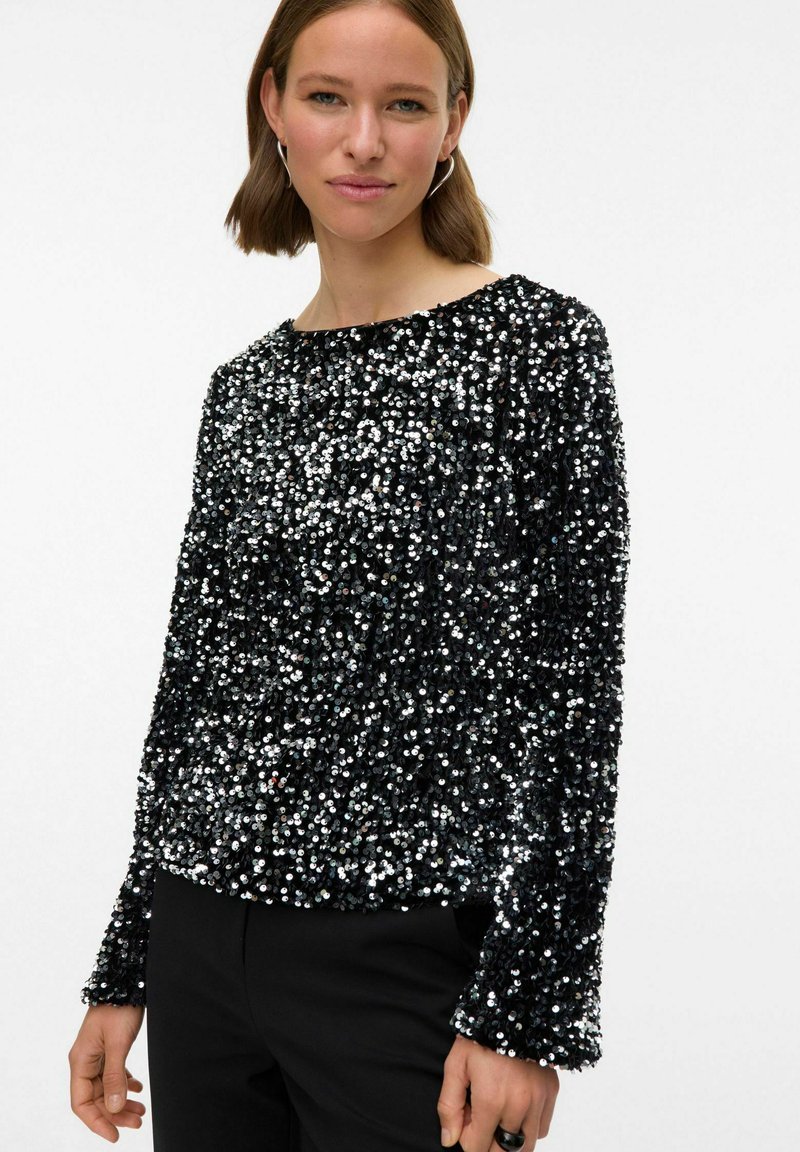 Črn zgornji del z bleščicami, ki ima okroglo izrezovanje, dolge rokave in teksturirano površino z odsevnimi bleščicami, v kombinaciji s črnimi hlačami.