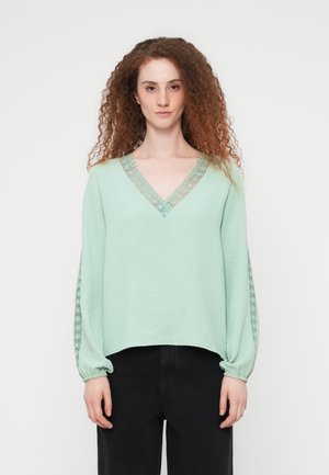 Γυναίκα με σγουρά κόκκινα μαλλιά, φοράει ανοιχτοπράσινο μπλουζάκι με δαντέλα στο V-λαιμό και τα μανίκια, σε συνδυασμό με μαύρο παντελόνι, στέκεται μπροστά σε λευκό φόντο.