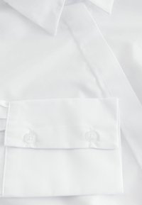Camicia bianca con colletto rigido e polsini con due bottoni trasparenti. Il tessuto appare liscio con una leggera lucentezza.