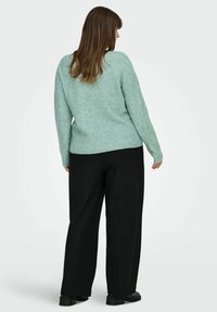 Šviesiai žalias megztinis iš mezgimo su tekstūruota medžiaga, plačiomis rankovėmis ir laisvo silueto, derinamas su aukštu liemeniu juodomis kelnėmis ir juodais batais.