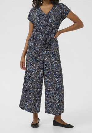 Kvinde iført en sort blomsterprint jumpsuit med korte ærmer og vide ben, kombineret med sorte balletsko, stående mod en ensfarvet baggrund.