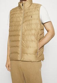 Beige puffer-väst med hög krage, framdragkedja och sidofickor. Har en kviltad design och en diskret logotyp på bröstet.