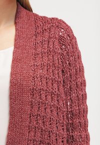 Gilet tricoté dans une couleur rose poussiéreux douce, avec un motif texturé en tissage ouvert et une coupe décontractée. Accentué par un large col côtelé.