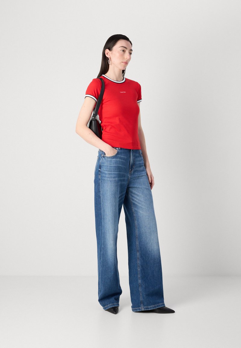 Camiseta roja de manga corta con ribete de rayas en blanco y negro, combinada con pantalones vaqueros de mezclilla azules de pierna ancha. La modelo lleva un bolso negro.