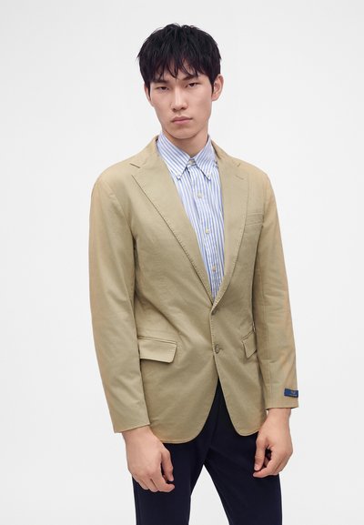 Ung mand iført en beige blazer over en blå- og hvidstribet skjorte med knapper og mørke bukser, stående mod en ensfarvet hvid baggrund.