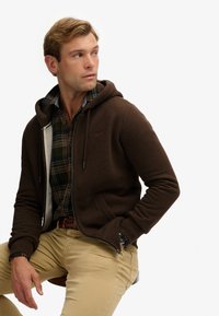 Hombre sentado en un taburete, vestido con una chaqueta con cremallera marrón, una camisa de cuadros verde y naranja, pantalones beige y un cinturón trenzado, mirando de lado.