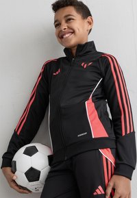 Smiling boy in black and red sportswear holds a soccer ball, leaning against a white wall.