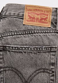 Černé džíny z denimové látky s béžovou koženou visačkou, na které je nápis "Levi Strauss & Co." a logo. Obsahují prošívané zadní kapsy a textured povrch.