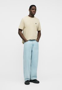 Άνδρας που φοράει μπεζ μπλουζάκι με μικρό λογότυπο, ανοιχτό μπλε τζιν παντελόνι φαρδιάς γραμμής, μαύρα αθλητικά παπούτσια και γυαλιά, στέκεται με τα χέρια στις τσέπες μπροστά σε λευκό φόντο.