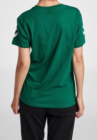 Camiseta de algodón verde con mangas cortas y cuello redondo. Presenta dos franjas blancas en cada hombro. Textura suave, ajuste holgado.