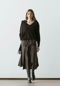 Brauner Strickpullover mit V-Ausschnitt, kombiniert mit einem braunen, plissierten Rock. Das Modell trägt schwarze kniehohe Stiefel und einen passenden Fransen-Schal.