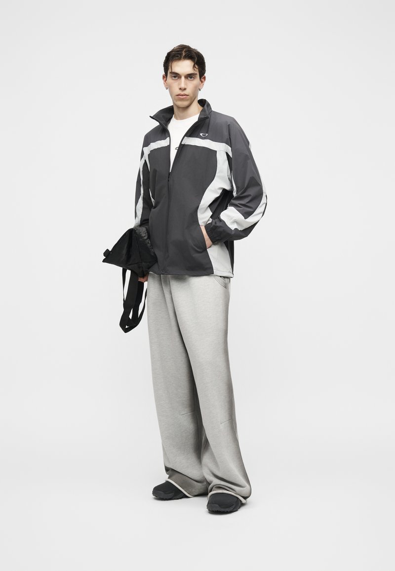 Joven con cortavientos negro y blanco, pantalones grises de pata ancha y zapatos negros, sosteniendo una bolsa negra a su lado.