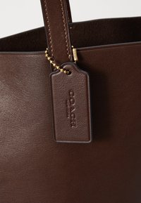 Borsa in pelle marrone con tracolla cucita e etichetta in pelle Coach New York attaccata da una piccola catena dorata.