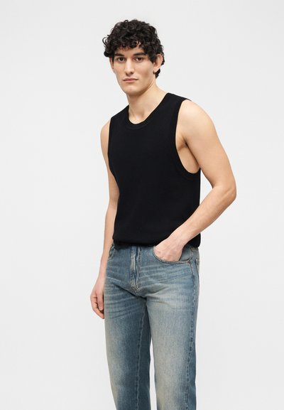 Νέος άνδρας με σγουρά μαλλιά, φορώντας μαύρο αμάνικο τοπ και ξεθωριασμένο μπλε τζιν, που στέκεται με το ένα χέρι στην τσέπη, μπροστά σε λευκό φόντο.