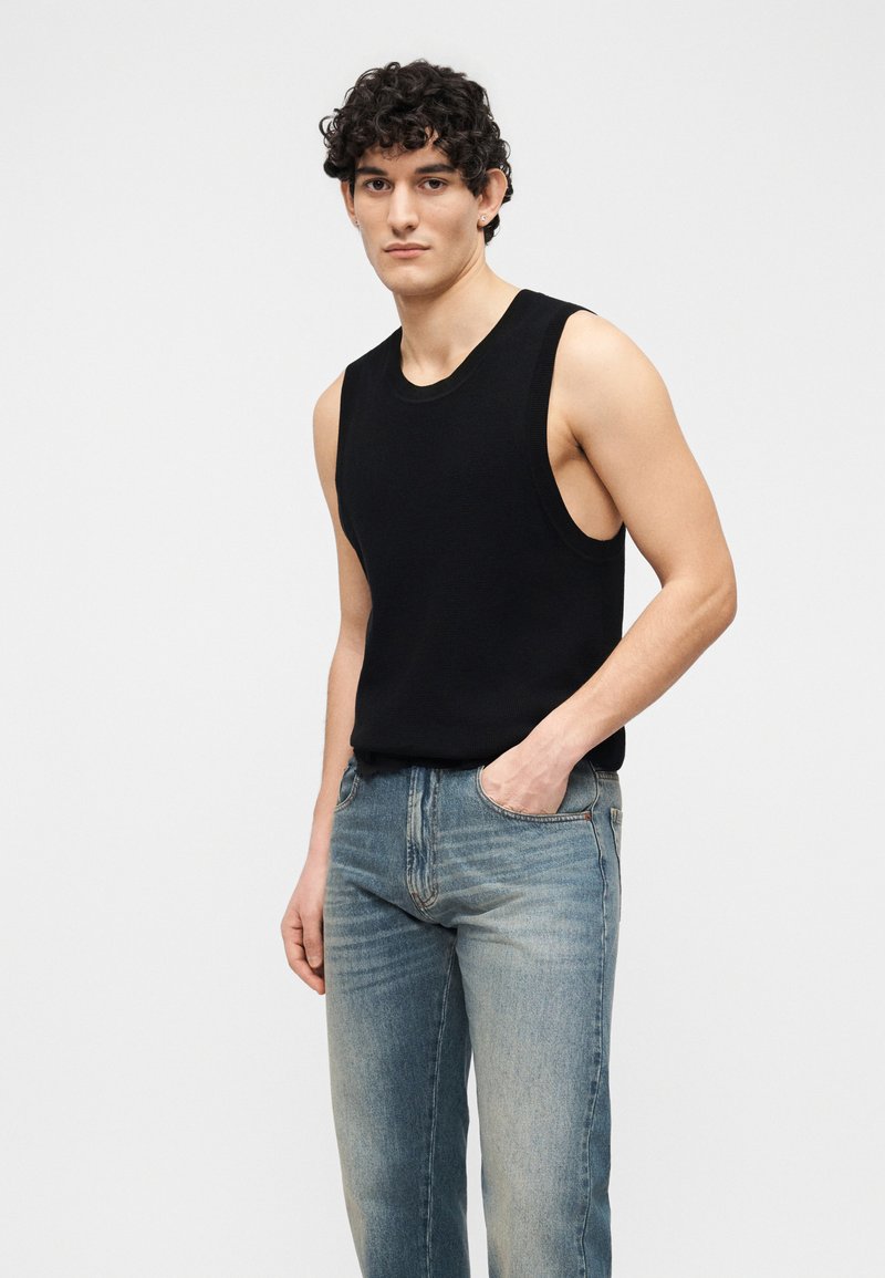 Jeune homme aux cheveux bouclés portant un débardeur noir et un jean bleu délavé, debout avec une main dans la poche, sur fond blanc.