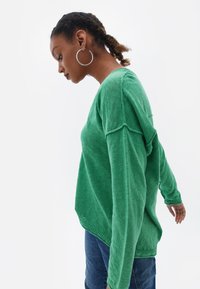 Maglione verde lavorato a maglia con maniche lunghe e vestibilità rilassata, caratterizzato da un collo rotondo e spacchi laterali, abbinato a jeans in denim blu.