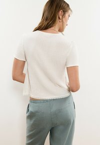 Mujer con cabello castaño claro lleva una blusa blanca de manga corta con textura y borde de encaje, junto con pantalones sueltos azul claro, de espaldas contra una pared lisa.