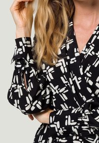 Blouse en tissu noir avec motif abstrait blanc, présentant un design enroulé et des manches bouffantes avec un accent en bouton et un détail découpé.