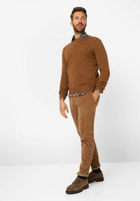 Brauner strukturierter Pullover über einem gemusterten blauen Hemd, kombiniert mit hellbraunen Cord-Hosen und braunen Wildleder-Doppelmönchschuhen.