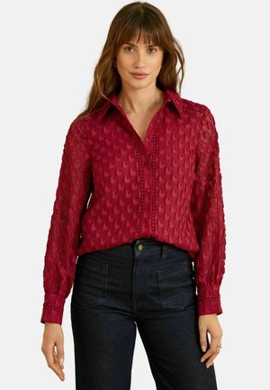 Rote transparente Bluse mit gestickten Herzmustern, durchgeknöpftem Vorderteil, Kragen und langen Ärmeln mit enganliegenden Bündchen. Kombiniert mit dunklem Denim.