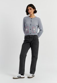 Model staat zelfverzekerd in een lichtgrijze cardigan met speelse zakontwerpen, gecombineerd met zwarte jeans en casual sneakers.