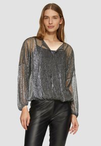 Blouse argentée transparente et scintillante avec des manches longues et un décolleté en V, associée à un pantalon en cuir noir ajusté. Matière légère et texturée.