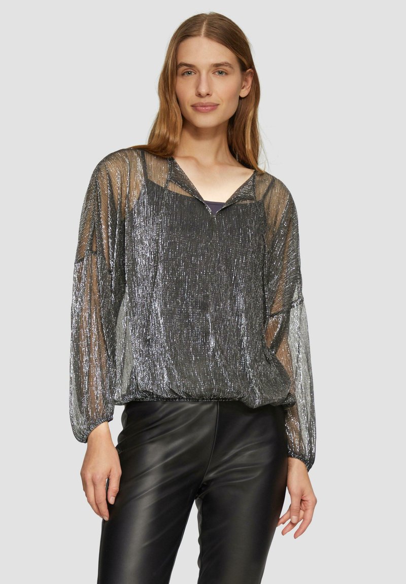 Blouse argentée transparente et scintillante avec des manches longues et un décolleté en V, associée à un pantalon en cuir noir ajusté. Matière légère et texturée.